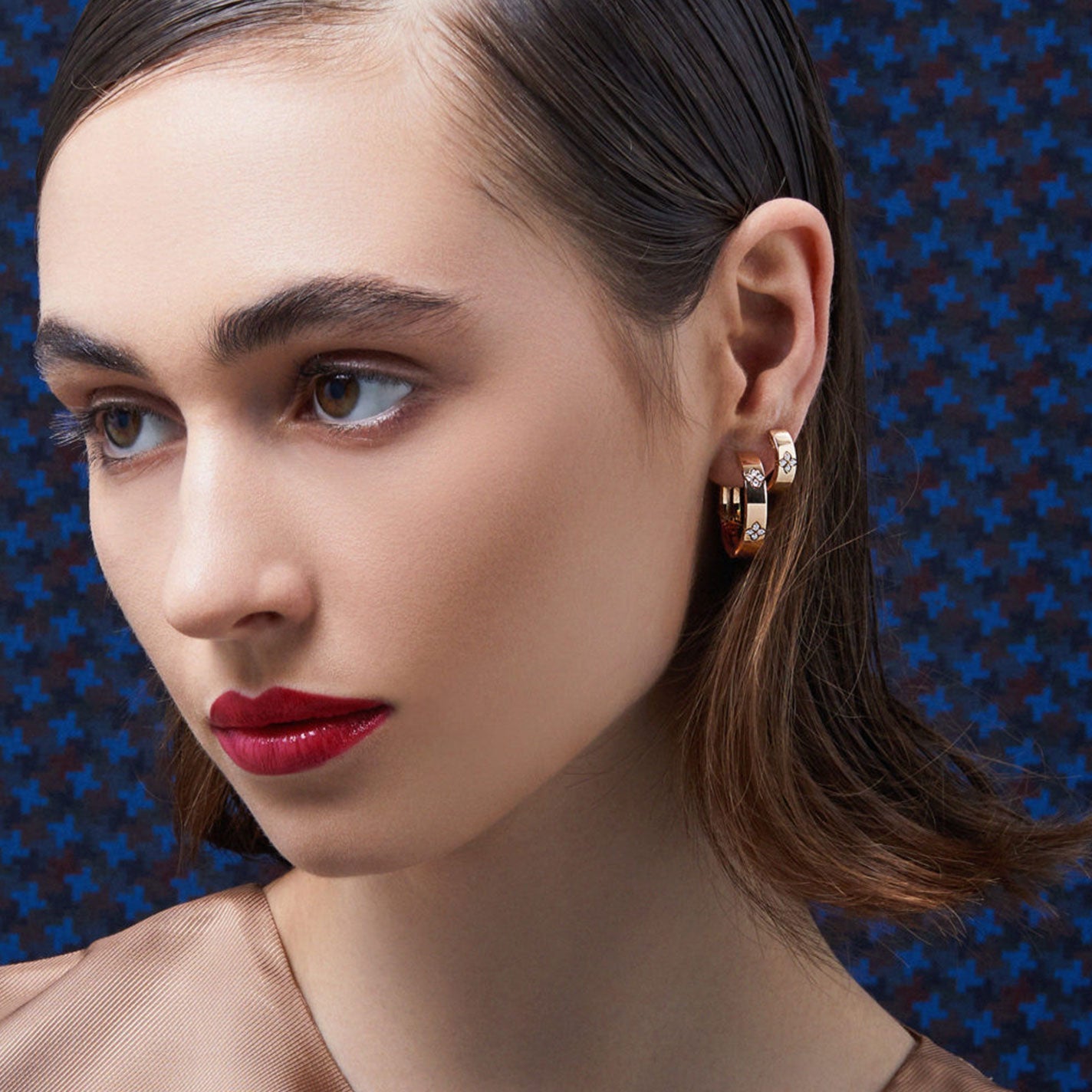 A woman with short brown hair and red lipstick gazes to the side. She is wearing Roberto Coin Love In Verona 18K Yellow Gold Diamond Huggie Earrings, perfectly complementing her sheer beige top. The background features a blue and black geometric pattern.