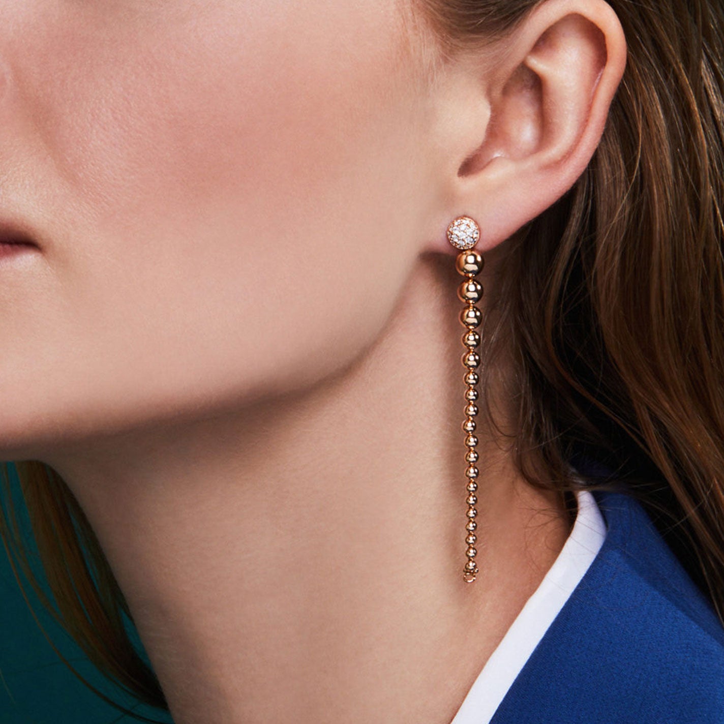 A close-up captures an individual adorned with Roberto Coin Tassel 18K Yellow Gold Diamond Convertible Drop Earrings, featuring a small diamond stud at the earlobe. The scene is set against a dark turquoise backdrop, highlighting the individual's light brown hair and blue blazer.