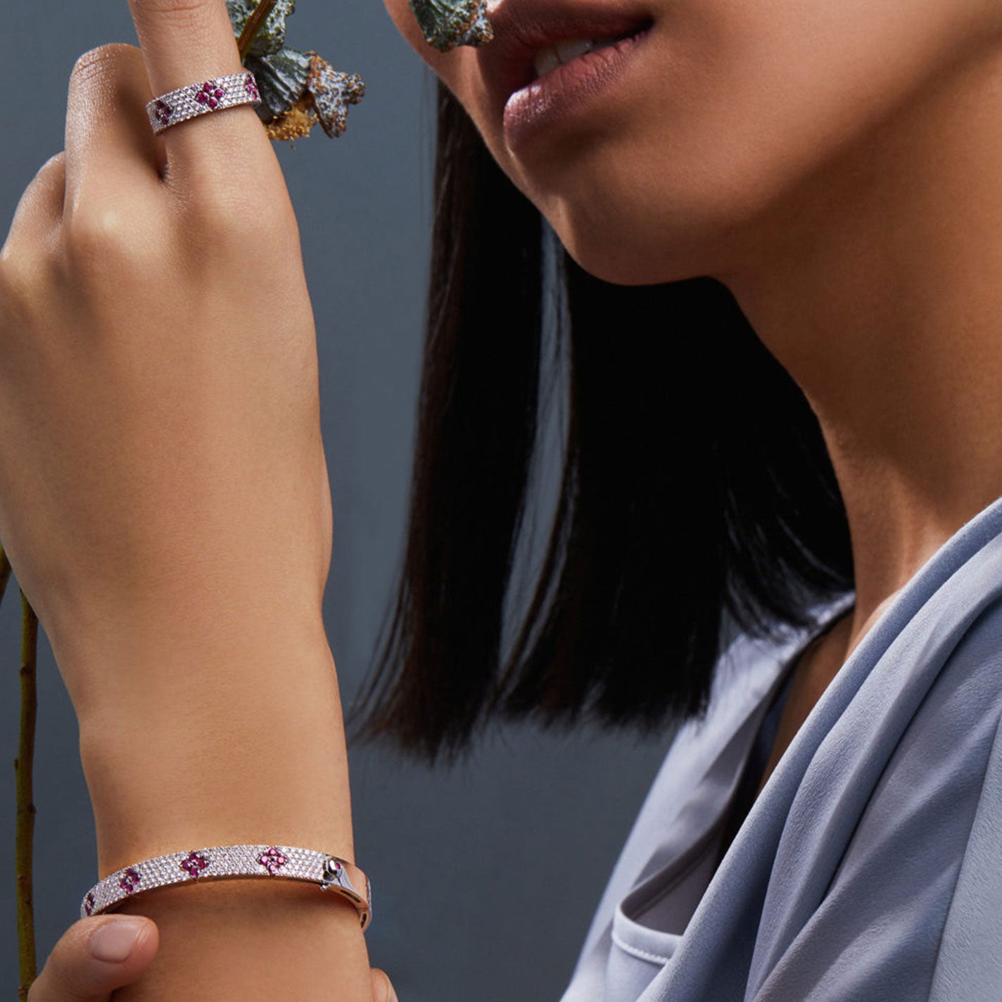 A woman with straight black hair gently holds a branch close to her face, showcasing a sparkling Roberto Coin Love In Verona 18K White Gold Diamond Bangle. She pairs it with a matching bracelet adorned with pink gemstone accents. Her attire is crafted from understated, light-colored fabric, set against a muted gray backdrop that exudes an elegant Love In Verona ambiance.