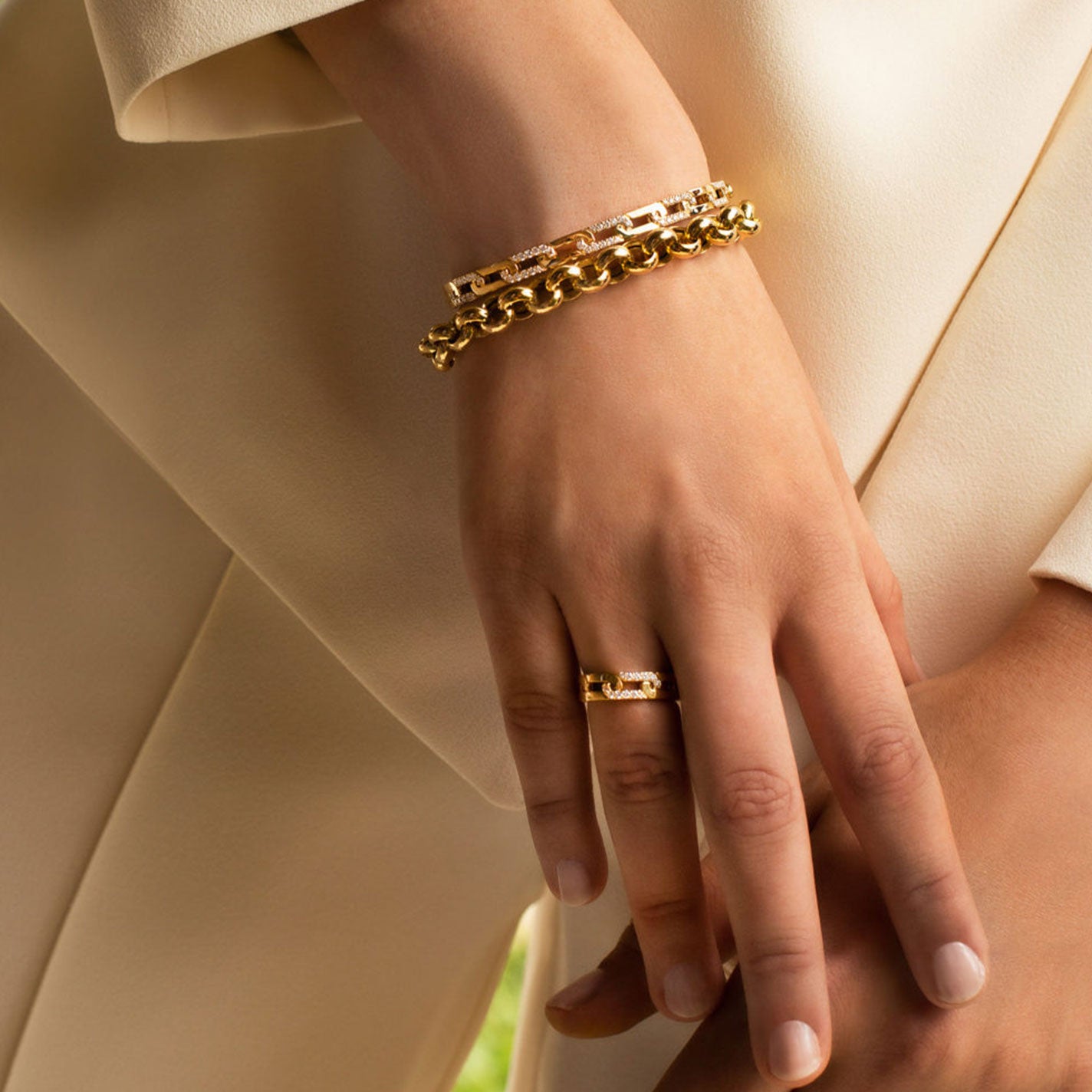 A close-up of a person's hand adorned with a Roberto Coin Navarra 18K Yellow Gold Diamond Accent Slim Ring and two gold chain bracelets. The individual is dressed in a light beige outfit, set against a softly blurred background.