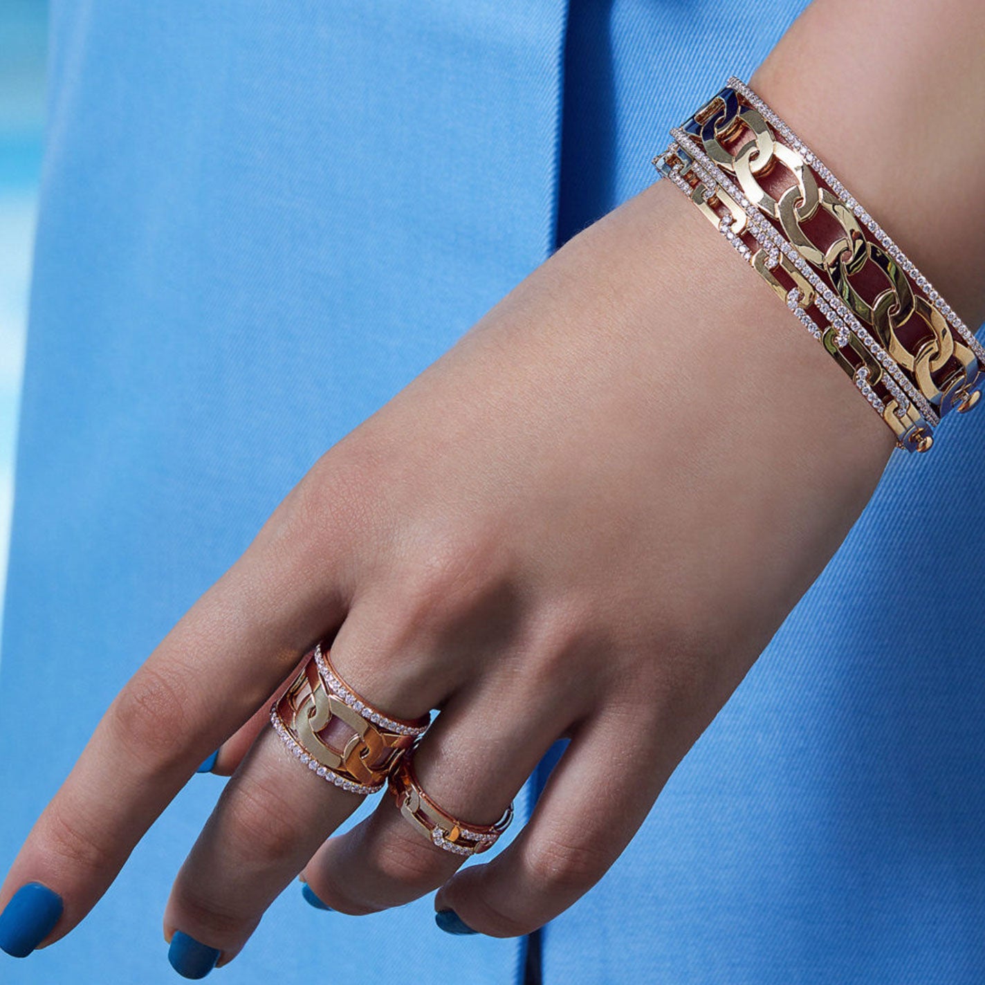 A person's hand showcases an exquisite mix of jewelry, featuring a Roberto Coin Navarra 18K Yellow Gold Diamond Wide Bangle alongside multiple rings crafted from rose gold and 18K yellow gold. Some of the pieces are elegantly studded with round diamonds. The hand rests gracefully against a light blue fabric background, with blue-painted nails that perfectly complement the attire.
