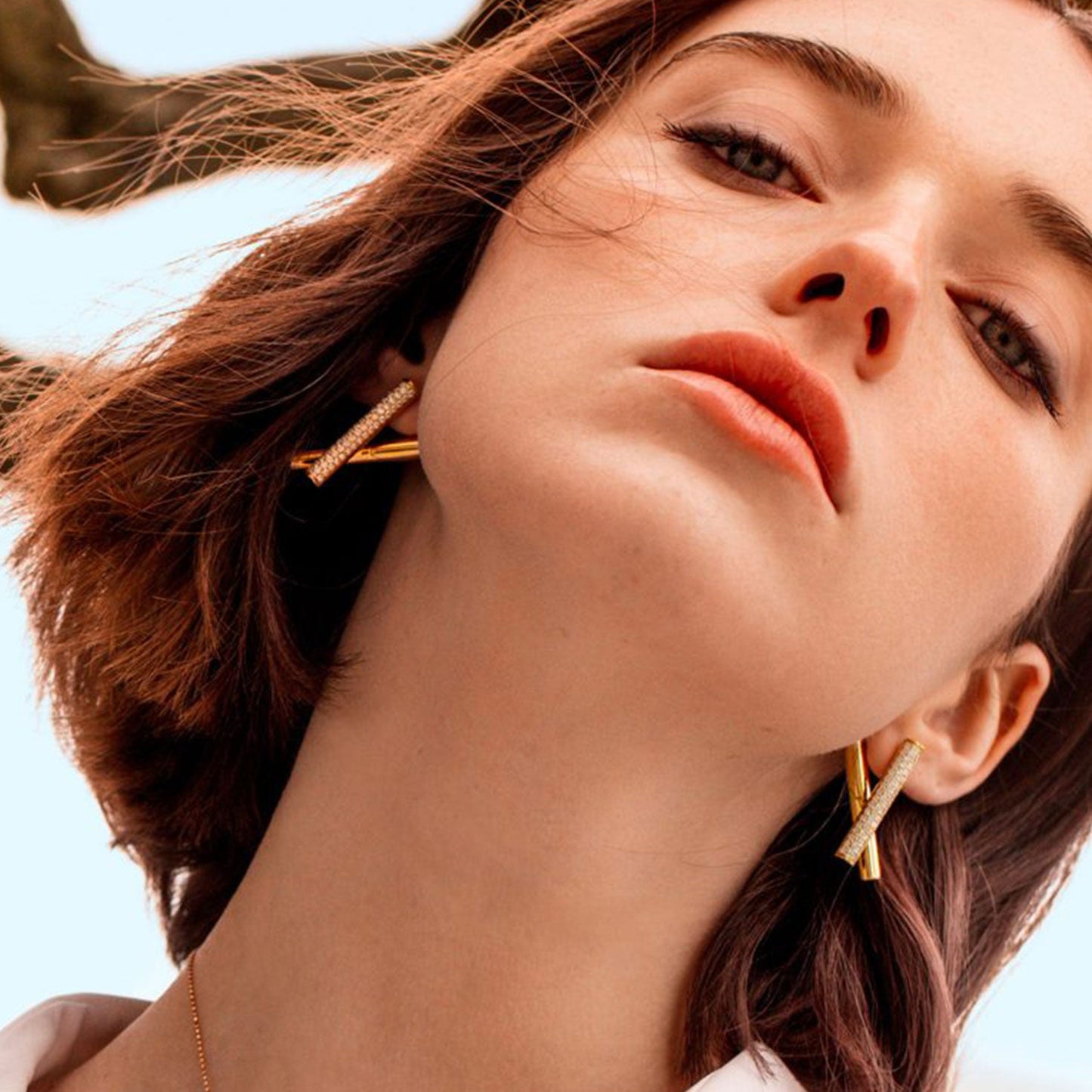 A woman with short brown hair gazes upward, wearing Roberto Coin Domino 18K Rose Gold Diamond Crossover Earrings and a white top. The background features blurred tree branches and a bright sky, highlighting her face and the elegant earrings.