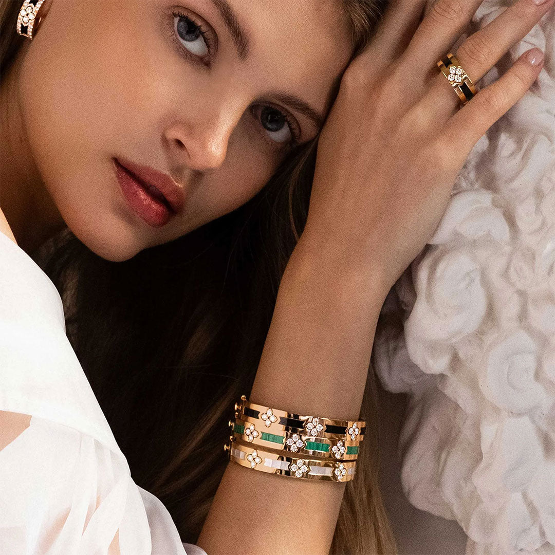 A woman rests her head on her arm, displaying a Roberto Coin Love In Verona 18K Yellow Gold Diamond with Malachite Bangle, beautifully crafted and adorned with floral designs. She complements it with a matching ring and wears a white top. Her expression is thoughtful against a softly lit background textured with white patterns.