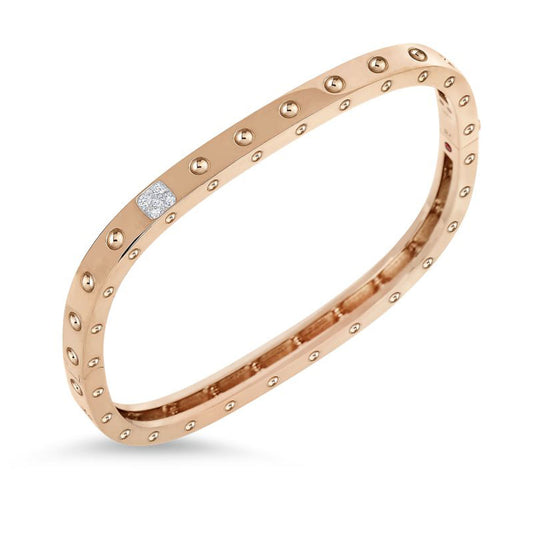 The Roberto Coin Pois Moi 18K Rose Gold Diamond Accent Square Bangle is a stunning piece that embodies modern sophistication through its sleek design. Crafted from 18K Rose Gold by renowned brand Roberto Coin, the bangle showcases small circular studs evenly distributed around its band and is highlighted by a single diamond set near the center, all elegantly presented against a plain white background.