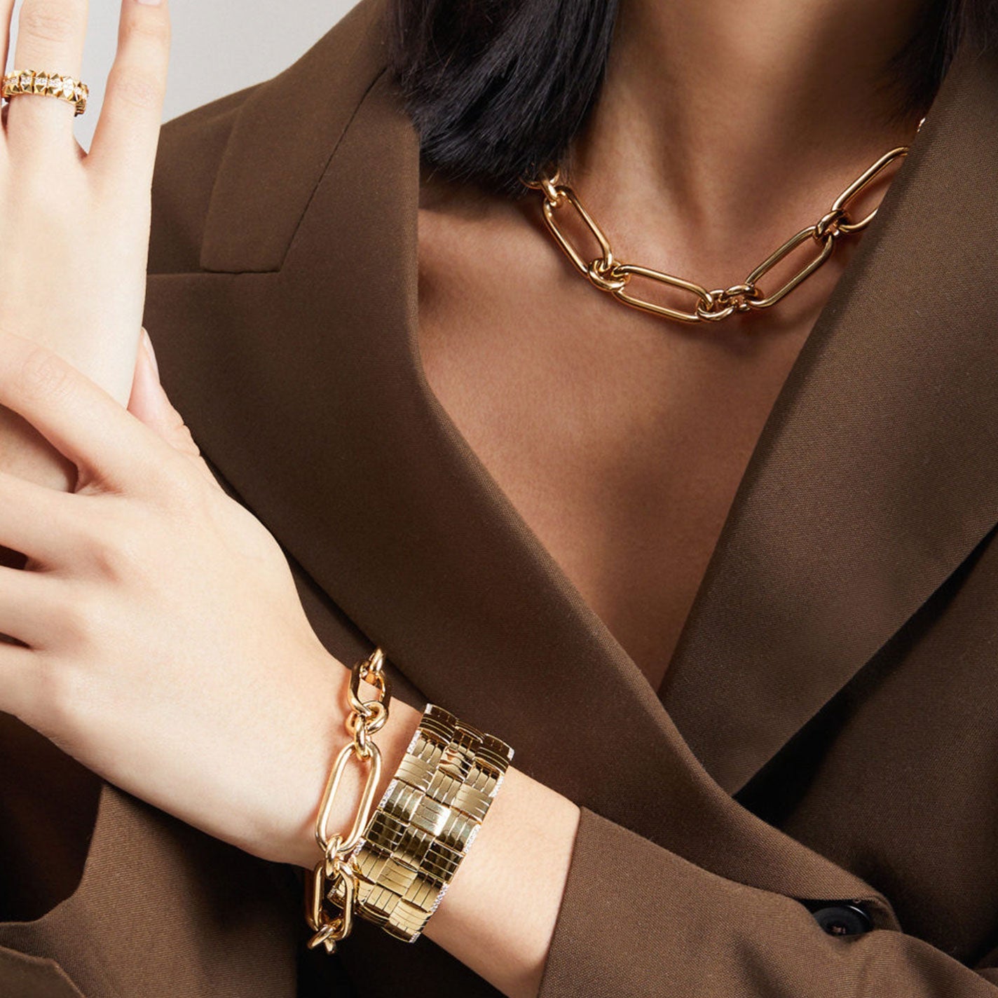 A person in a brown blazer showcases luxurious gold jewelry, highlighting the Roberto Coin 18K Rose Gold Bracelet. A hand rests on the chest, adorned with an exquisite 18 Karat Rosé Gold ring featuring small stones. The impeccable craftsmanship emphasizes elegance and refinement.