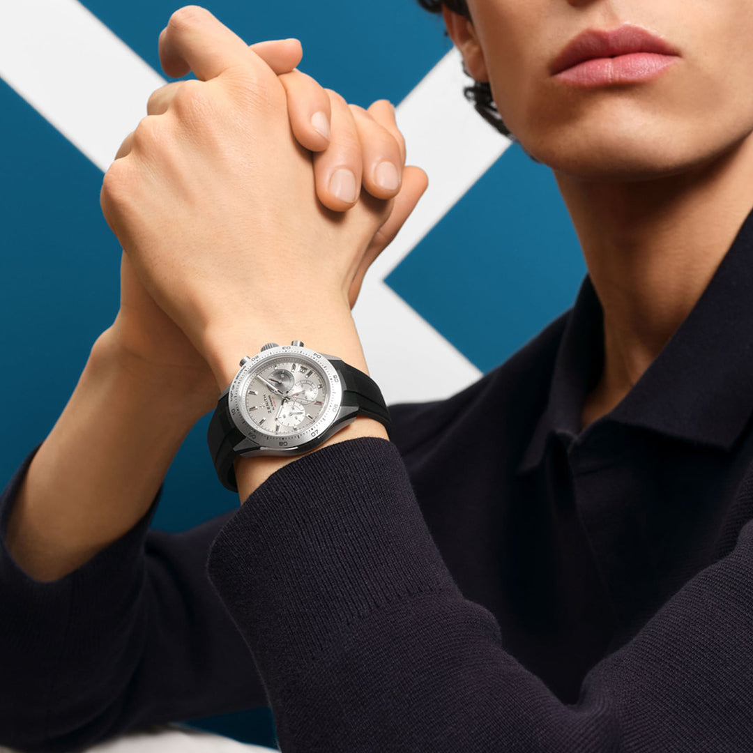 A person in a black long-sleeve shirt rests their chin on clasped hands, wearing a sophisticated Zenith 41mm Chronomaster Sport watch with a black strap. The timepiece features the El Primero 3600 movement in a titanium case, standing out against the geometric blue and white background.