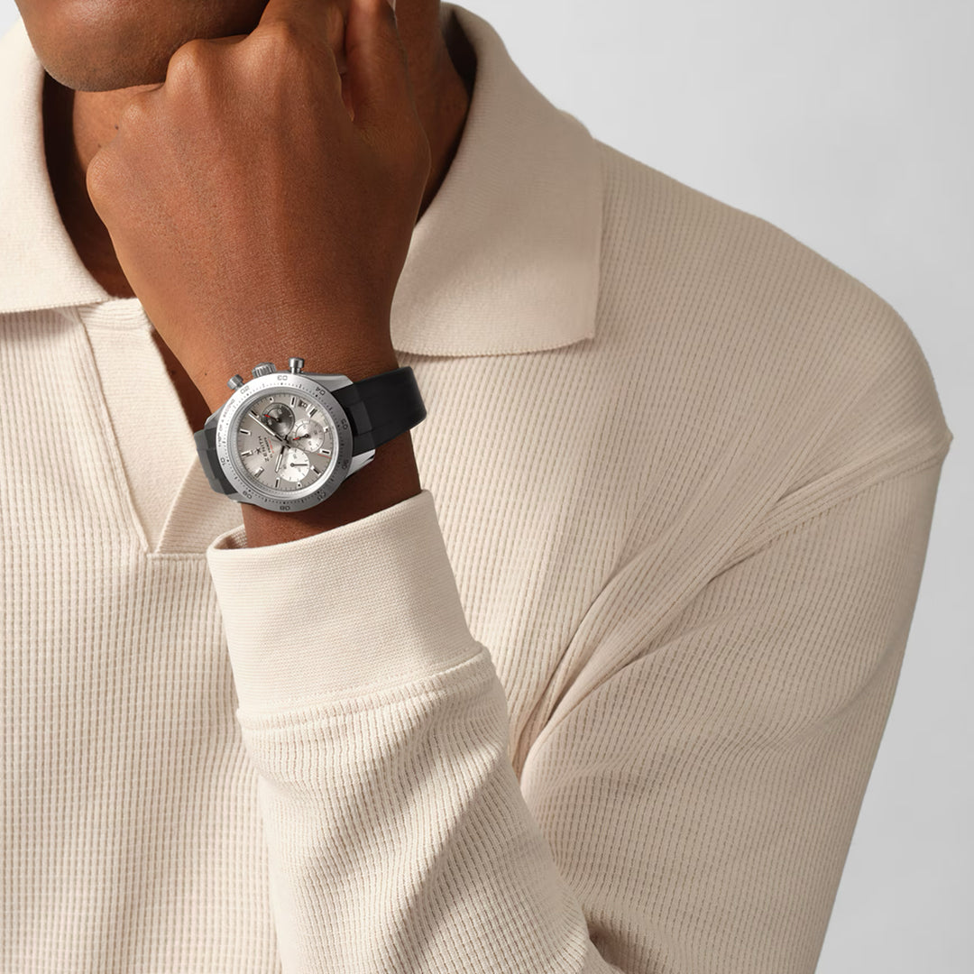 A person in a beige ribbed sweater rests their chin on their hand, showcasing the Zenith ZENITH Chronomaster Sport El Primero 3600 41mm watch with a black strap. The titanium case subtly gleams against the plain white background.