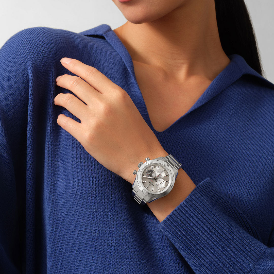 A woman in a blue sweater elegantly showcases the ZENITH Chronomaster Sport, featuring the El Primero 3600 movement and three sub-dials, with its detailed round face and titanium case. Her arm is bent at the elbow, hand gently resting on her shoulder.