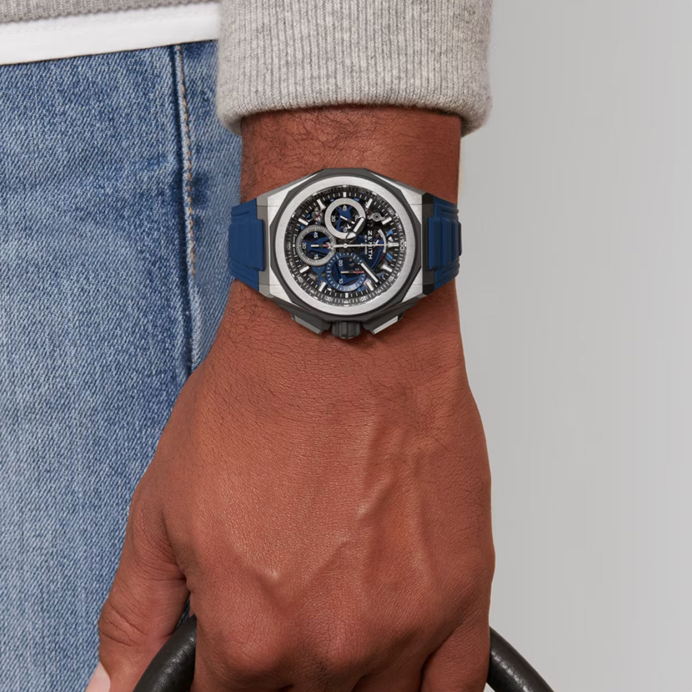 Close-up of a person's wrist adorned with a sleek ZENITH Defy Extreme El Primero 21 Automatic Chronograph 45mm Watch. The sophisticated dial and blue strap accentuate the timepiece, glimmering as the person grips a curved, black object. Set against a neutral, light background that complements their casual attire.