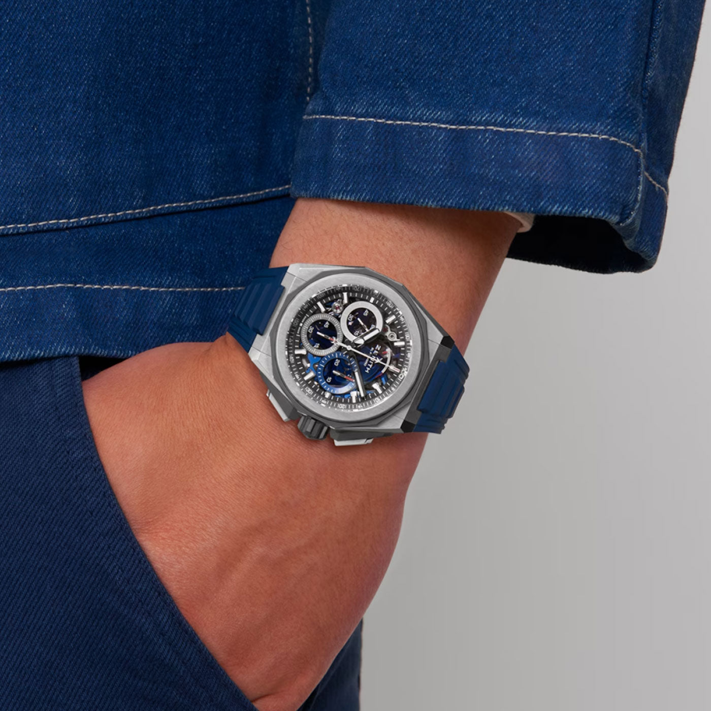 A person is wearing a blue denim jacket with their hand in their pocket, showcasing the ZENITH Defy Extreme El Primero 21 Automatic Chronograph 45mm Watch on their wrist. This timepiece is made of titanium, featuring a silver finish and an intricate blue and black dial, which is perfectly complemented by its matching dark blue strap from Zenith.