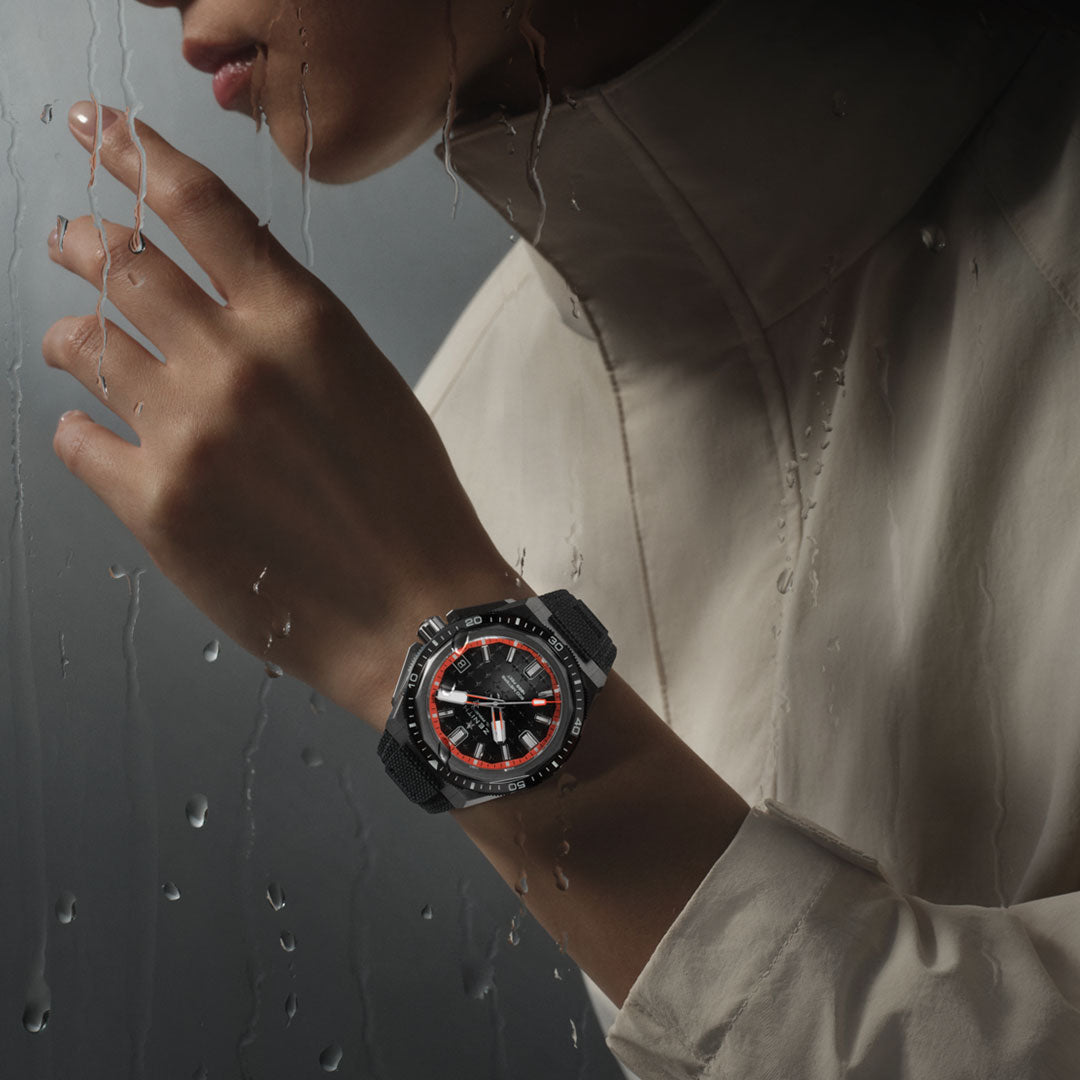 A person in a light-colored coat with the hood up stands by a rain-soaked glass window, their ZENITH Defy Extreme Diver El Primero 3620 Automatic Chronograph wristwatch gleaming. The titanium elements of this 42.5mm Zenith watch add to the moody, atmospheric scene.