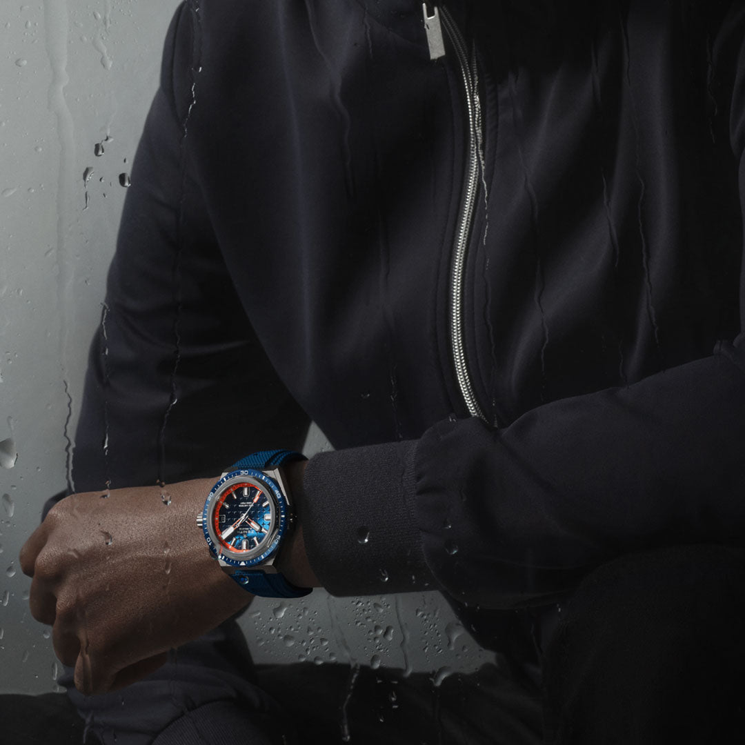 A person in a dark jacket sits by a rain-speckled window, donning the ZENITH Defy Extreme Diver El Primero 3620 Automatic Chronograph 42.5mm Watch on their wrist. The watch features an orange-and-blue face set in a sleek titanium case, crafted with Zenith's signature automatic movement. The dimly lit scene evokes a reflective mood.