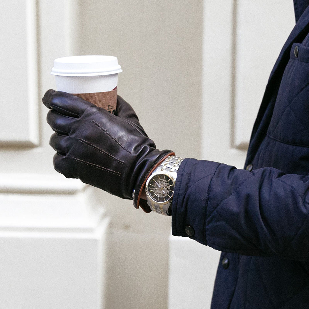 A person in a navy jacket and brown leather gloves holds a white disposable coffee cup. On their left wrist, they wear the Bulova Sutton Automatic 43mm Watch with a skeletonized dial. The background consists of cream-colored walls.