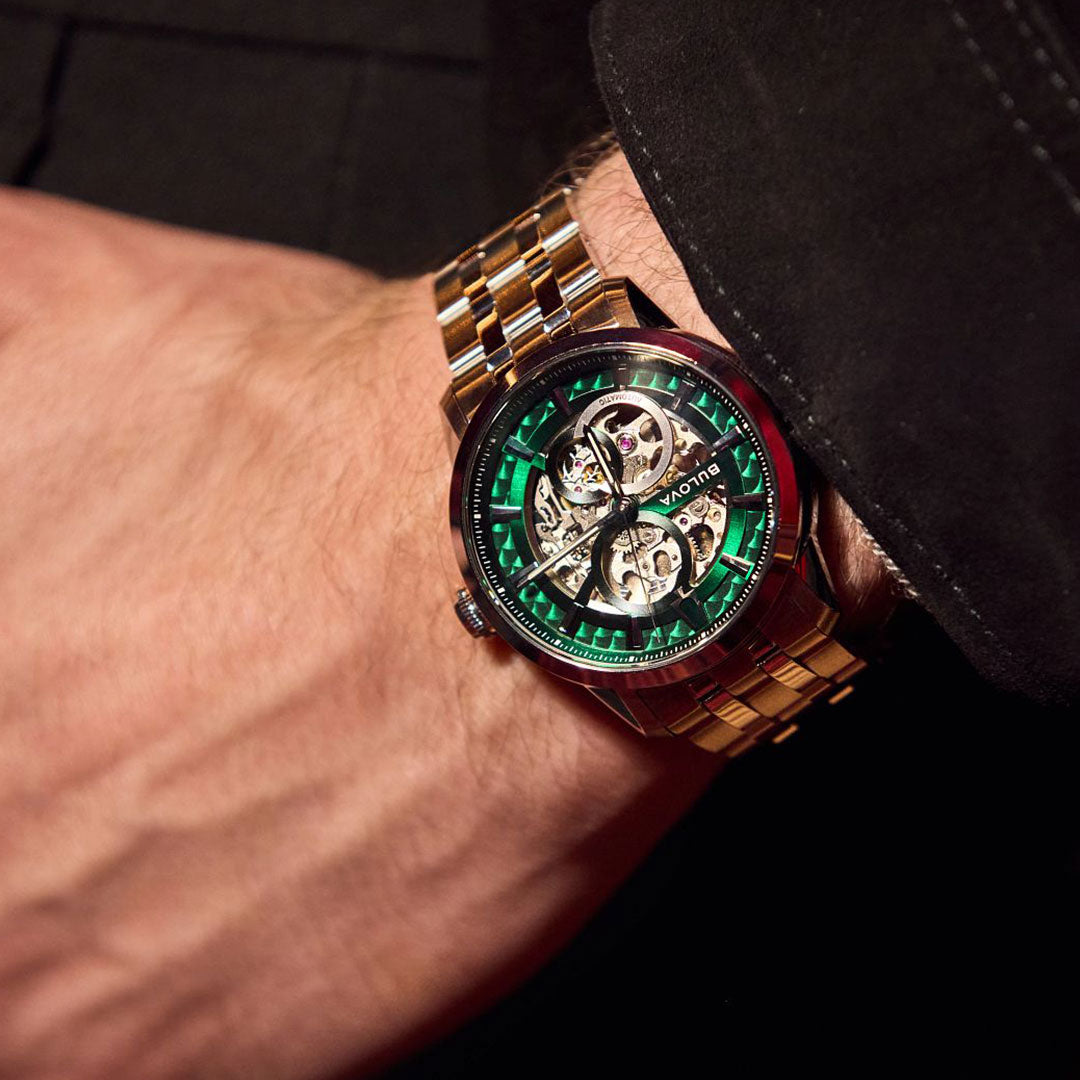 A close-up of a wrist wearing the Bulova Sutton Classic Automatic 40mm Watch with a green skeleton dial and stainless steel bracelet, showcasing intricate gears against a dark background.