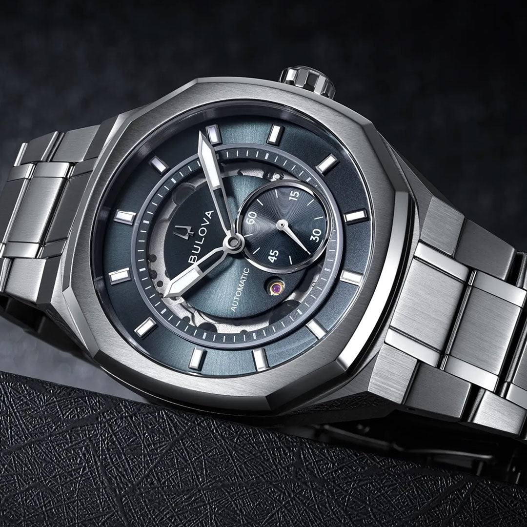 A close-up of the Bulova Surveyor Marc Anthony Automatic 40.5mm men’s watch showcases its silver metal band, dark dial with a visible subdial, and automatic movement, elegantly displayed on a textured black surface.