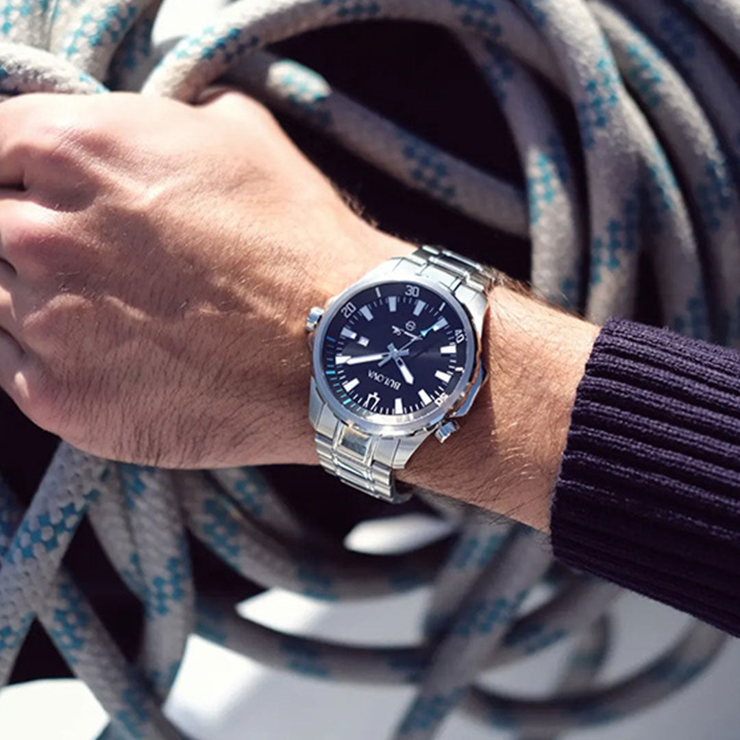A person wearing a Bulova Marine Star Series B Quartz 44mm Watch by Bulova, featuring a silver band and black face, rests their hand on coiled rope. The person is dressed in a dark, ribbed sleeve.