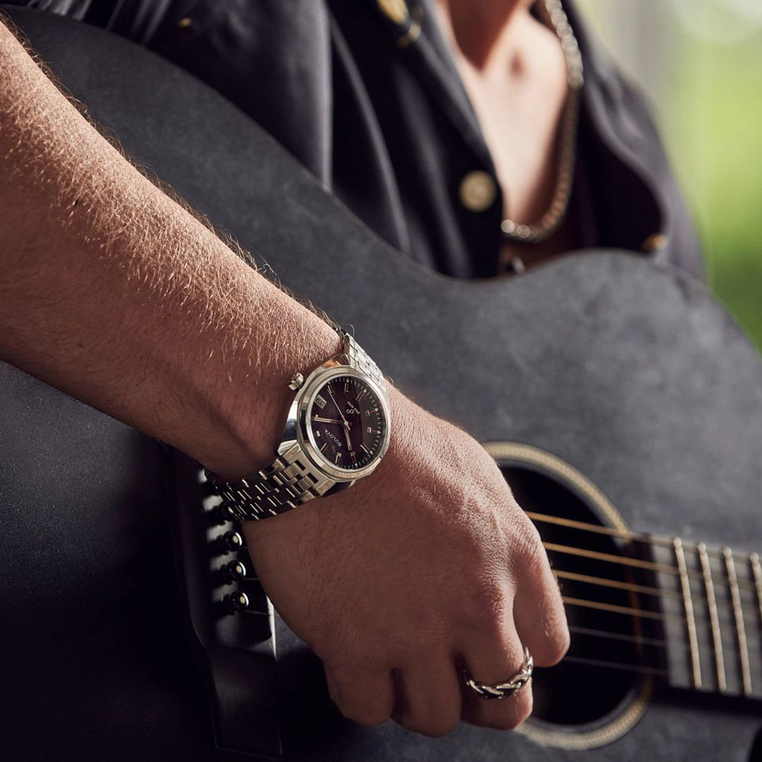 A person wearing a Bulova Jet Star Quartz 40mm watch with a chain bracelet plays an acoustic guitar. They are dressed in a black shirt with colorful lining, and the background is softly blurred.