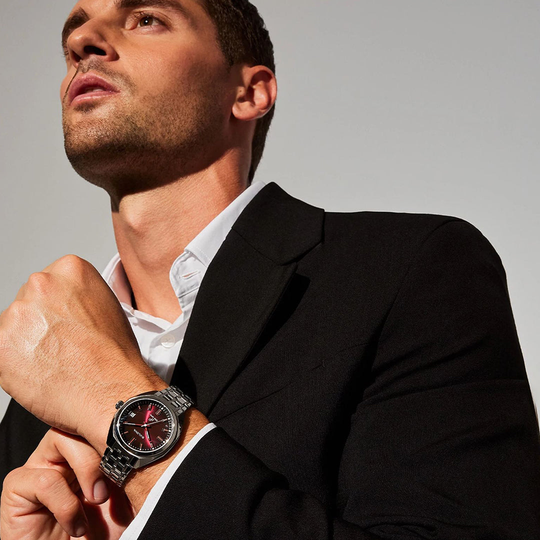 A man in a black suit looks up, displaying a Bulova Jet Star Quartz 40mm Watch with a sophisticated stainless steel case and striking red face on his left wrist. He pairs it with a white dress shirt under the suit, contrasted against a simple, light-colored background.