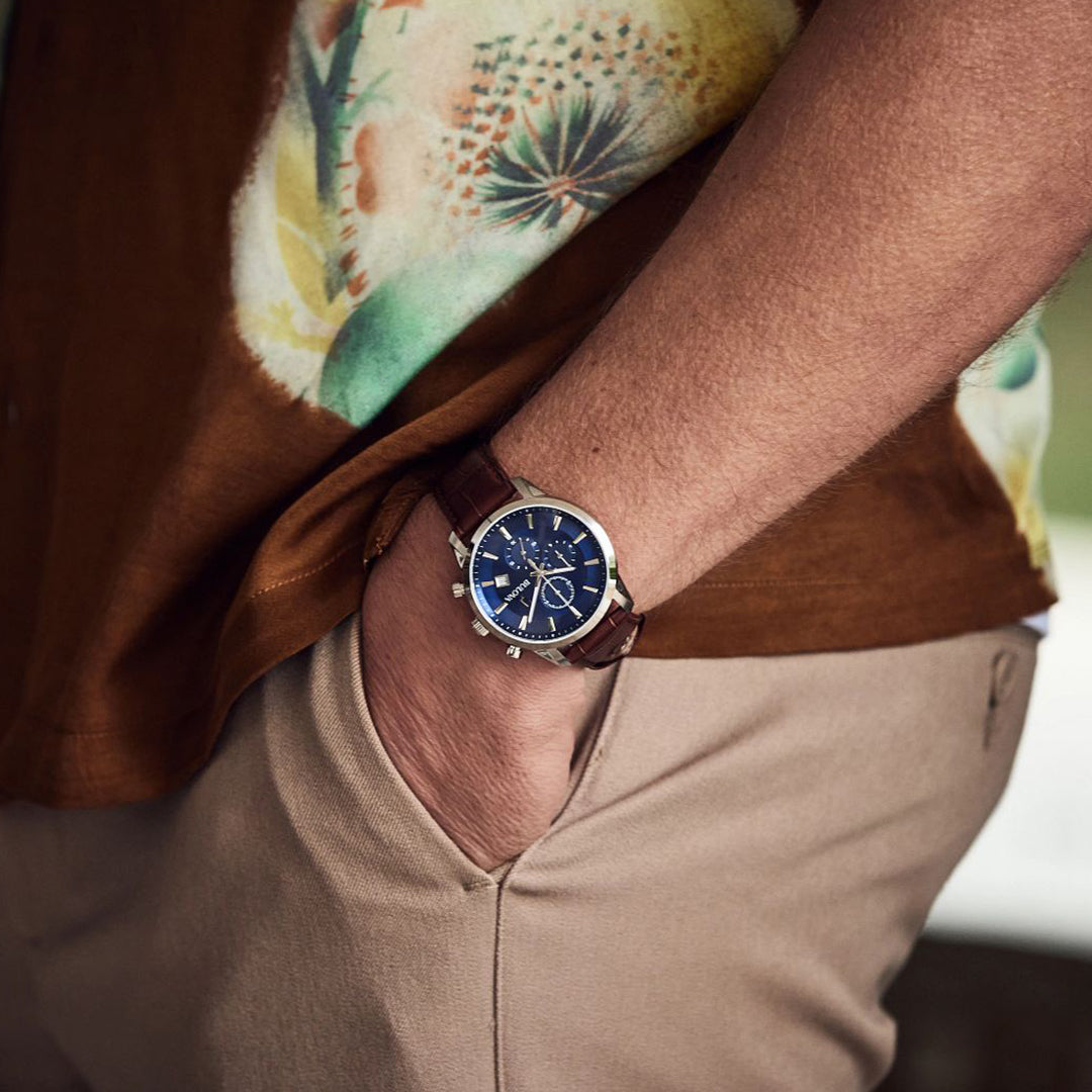 A person wearing a brown floral short-sleeve shirt and beige pants has their hand in their pocket, showcasing the Bulova Sutton Quartz 41mm Watch. The blue dial seamlessly complements the silver-tone stainless steel case and brown leather strap, enhancing their outfit.