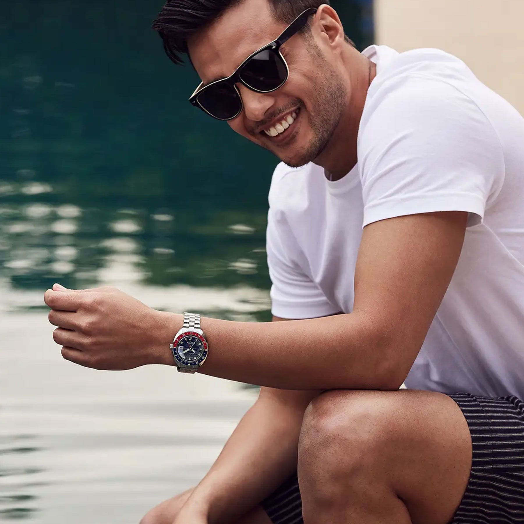 A smiling individual wearing sunglasses, a white t-shirt, and black shorts sits by the water, showcasing the Bulova Oceanographer Automatic GMT 41mm Watch on their left wrist. The watch's luminescent hour markers glint in the light, while the calm body of water in the background adds to the relaxed atmosphere.