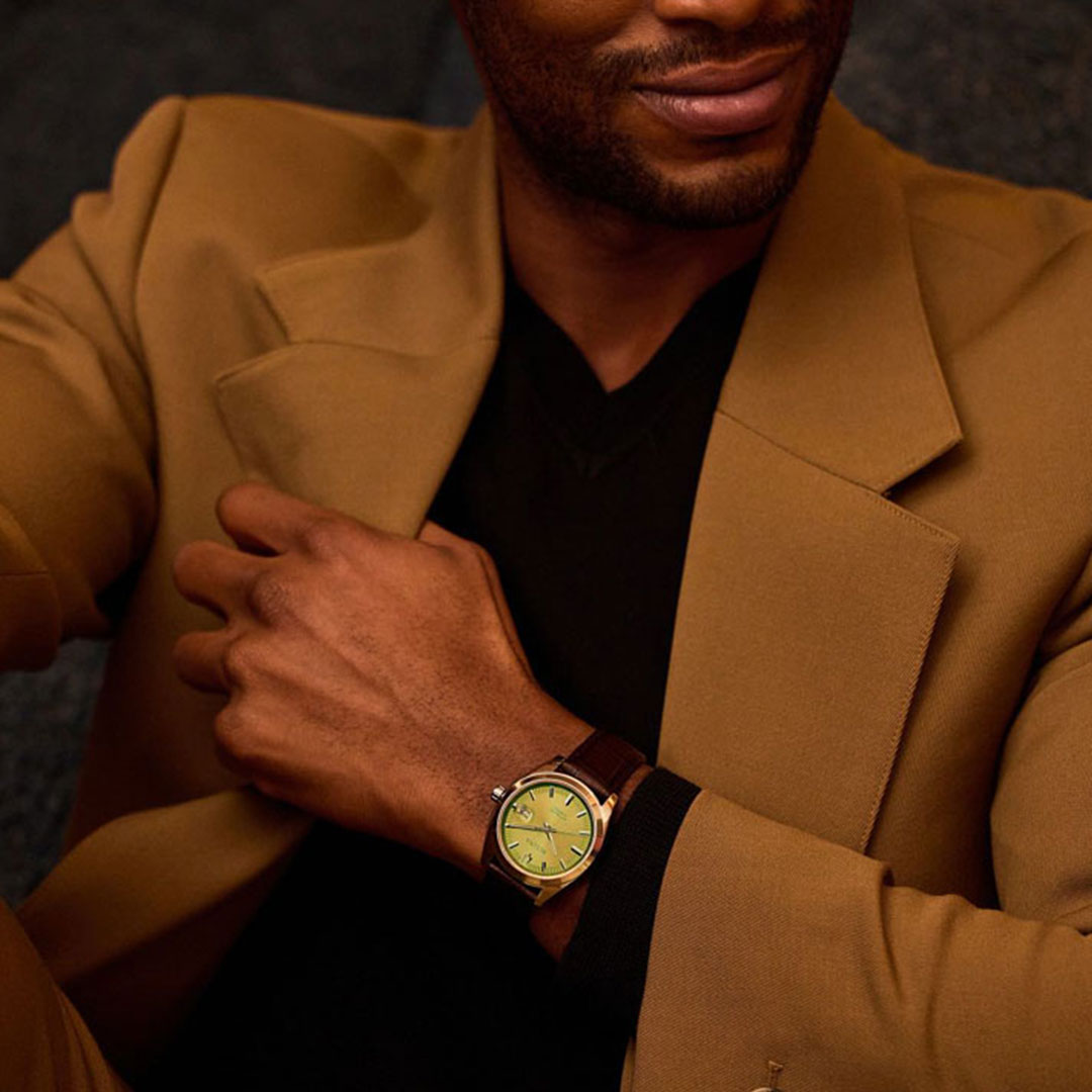 A person dressed in a tan suit and black shirt showcases the Bulova Surveyor Automatic 39mm Watch, which boasts a green face, brown leather strap, and stainless steel case. The elegant design of this Bulova timepiece, featuring an automatic 21-jewels movement, perfectly complements the attire.