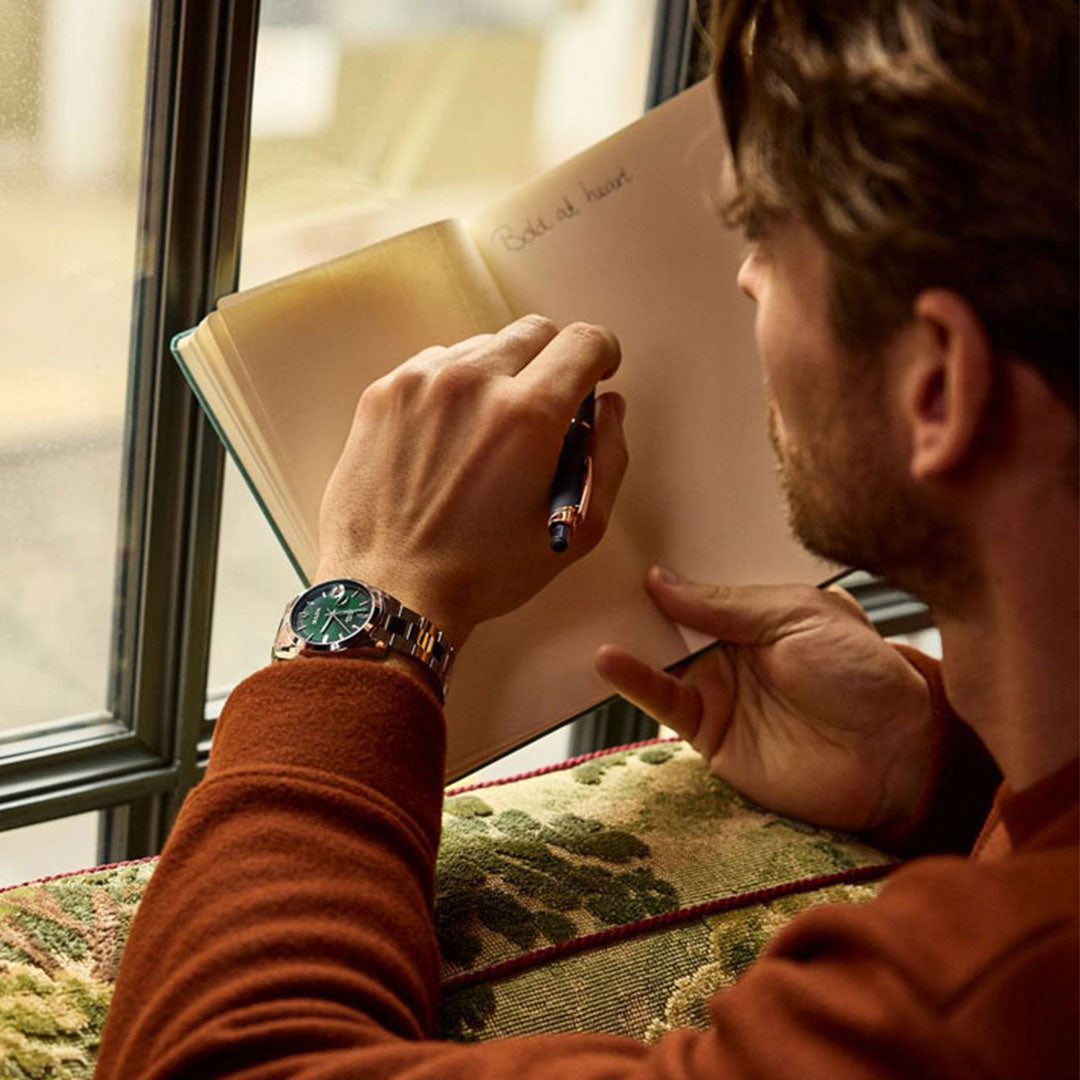 A person wearing an orange sweater and a gleaming Bulova Surveyor Automatic 39mm Watch featuring a stainless steel bracelet is writing with a pen in a notebook beside a window. The open page of the notebook displays "Gold at heart.