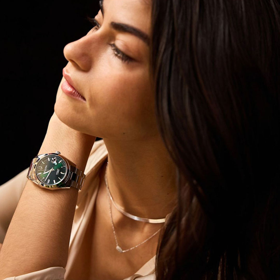 A woman with long dark hair gazes to the side, her wrist adorned with a Bulova Surveyor Automatic 39mm Watch, featuring a green face. Her simple necklace stands out against the dark background, elegantly highlighting her stainless steel bracelet and other accessories.