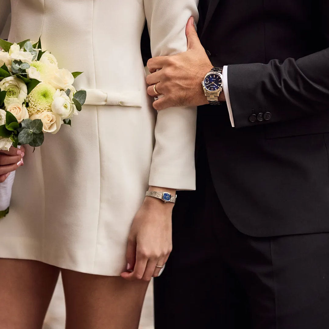 A couple dressed formally, with the woman in a white long-sleeve dress holding pale yellow and white flowers, and the man in a black suit wearing a Bulova Surveyor Automatic 39mm Watch from Bulova with a stainless steel bracelet.