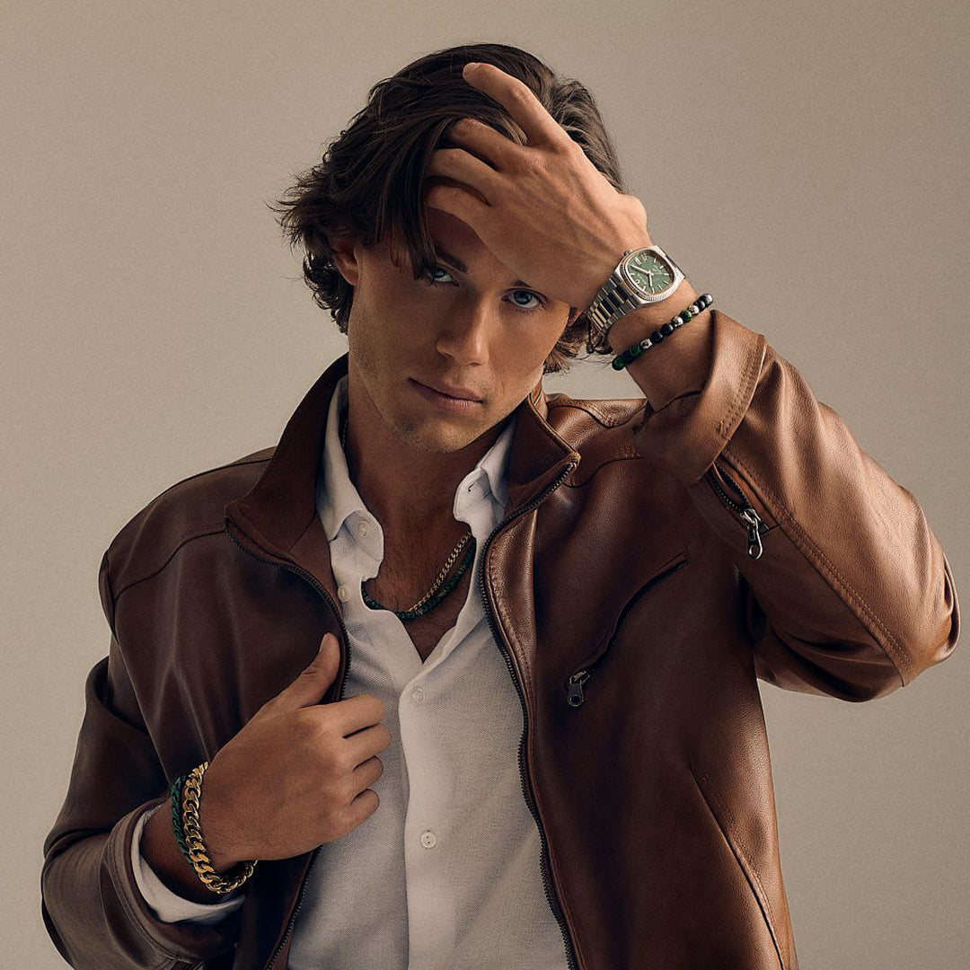 Wearing a brown leather jacket over a white shirt, a person with medium-length hair touches their head, clutching the jacket. They pose confidently against a plain background, showcasing a Bulova Super Seville HPQ Precisionist Quartz 37.5mm Watch with a stainless steel bracelet.