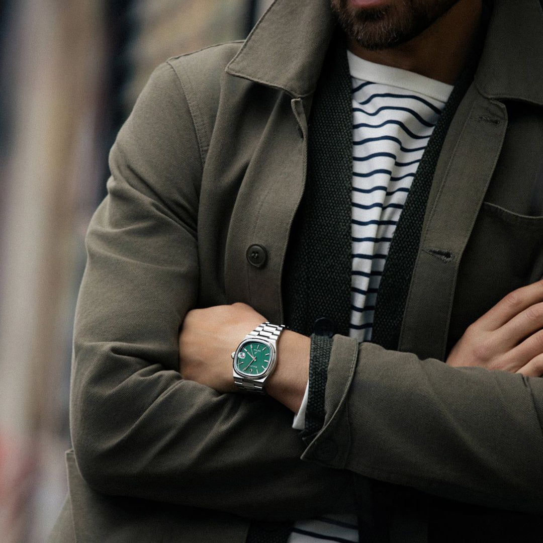 An individual in a green coat and striped shirt crosses their arms, displaying a Bulova Super Seville HPQ Precisionist Quartz 37.5mm watch with a green face and stainless steel bracelet. The background is blurred, emphasizing the Precisionist technology and stylish attire.