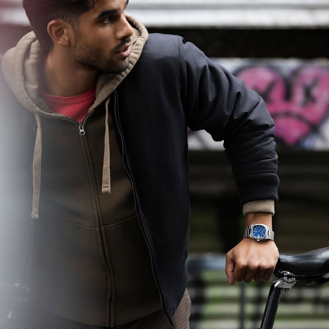 A person in a black jacket and brown hoodie casually leans on a bike, showcasing the Bulova Super Seville HPQ Precisionist Quartz 37.5mm Watch with its striking blue face. The stainless steel bracelet catches the vibrant pink and black graffiti art that decorates the background.