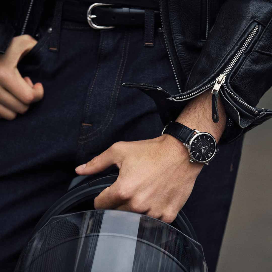 A person dressed in a black leather jacket and dark jeans holds a motorcycle helmet in one hand while adjusting their belt with the other. On their wrist glints the Bulova Hudson Automatic 39mm Watch, its silver casing housing an elegant dress watch with automatic movement.