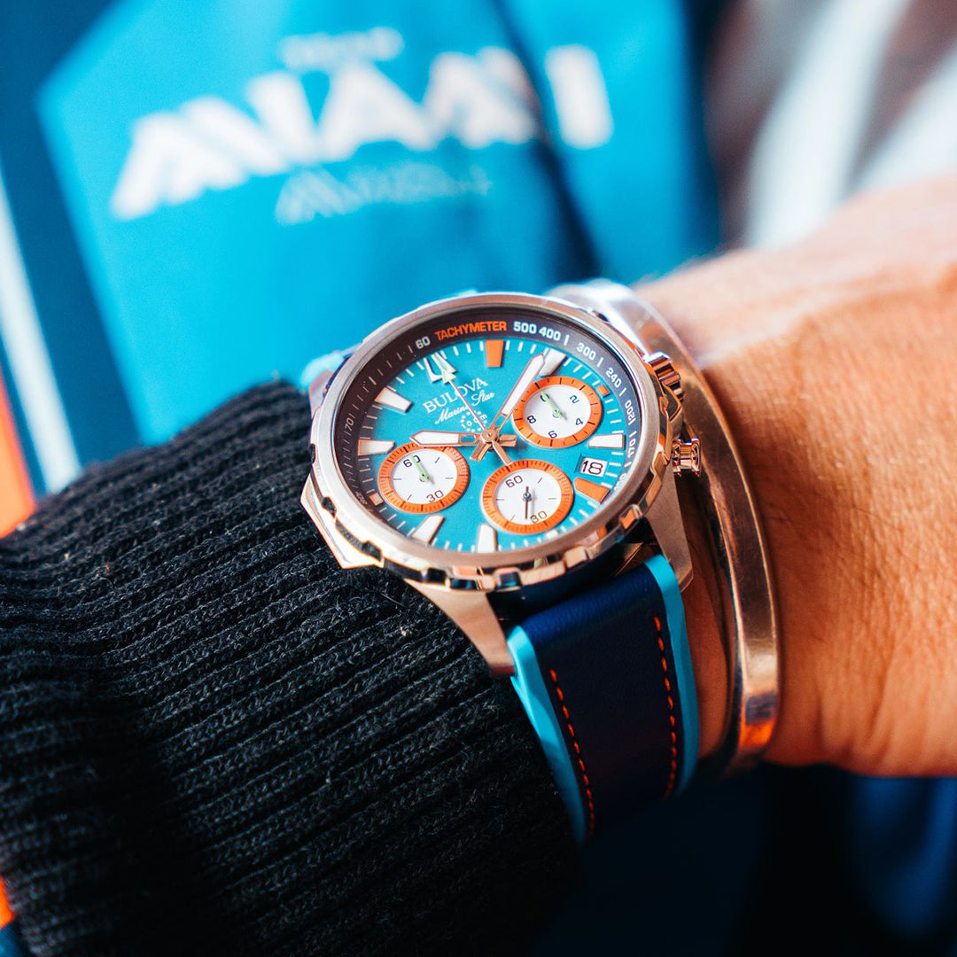 A wrist displays the Bulova E1 Team Miami Quartz 43mm Watch with a blue and orange face, celebrating Team Miami E1's racing, paired with sleek silver bracelets. The vibrant design pops against a blurred blue and orange backdrop, showcasing its unique charm.