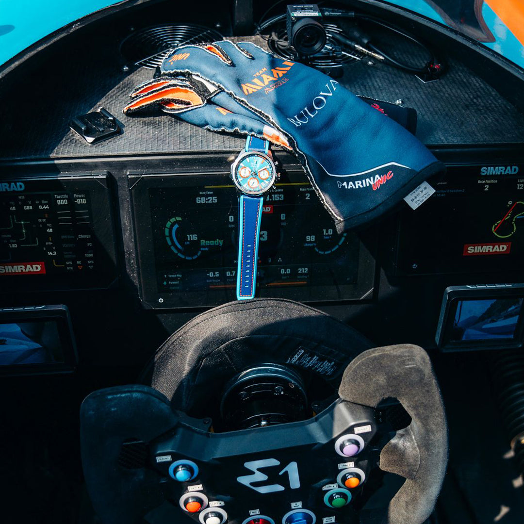 A close-up of a race car's cockpit shows a steering wheel with colorful buttons, along with racing gloves and the Bulova E1 Team Miami Quartz 43mm Watch on the dashboard, surrounded by digital displays.