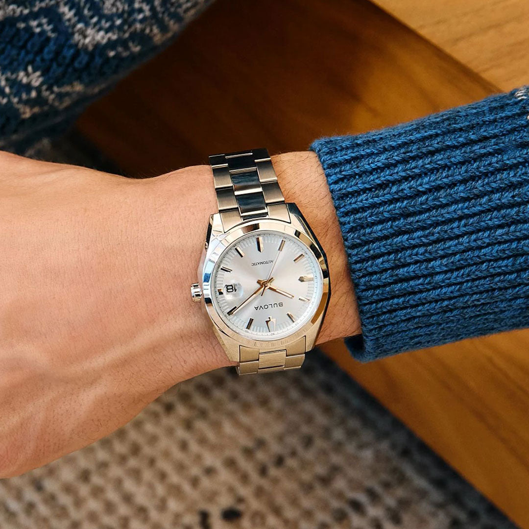Wearing a Bulova Surveyor Automatic 39mm Watch with a stainless steel band and white dial, a person in a blue knit sweater rests their arm on a wooden surface.