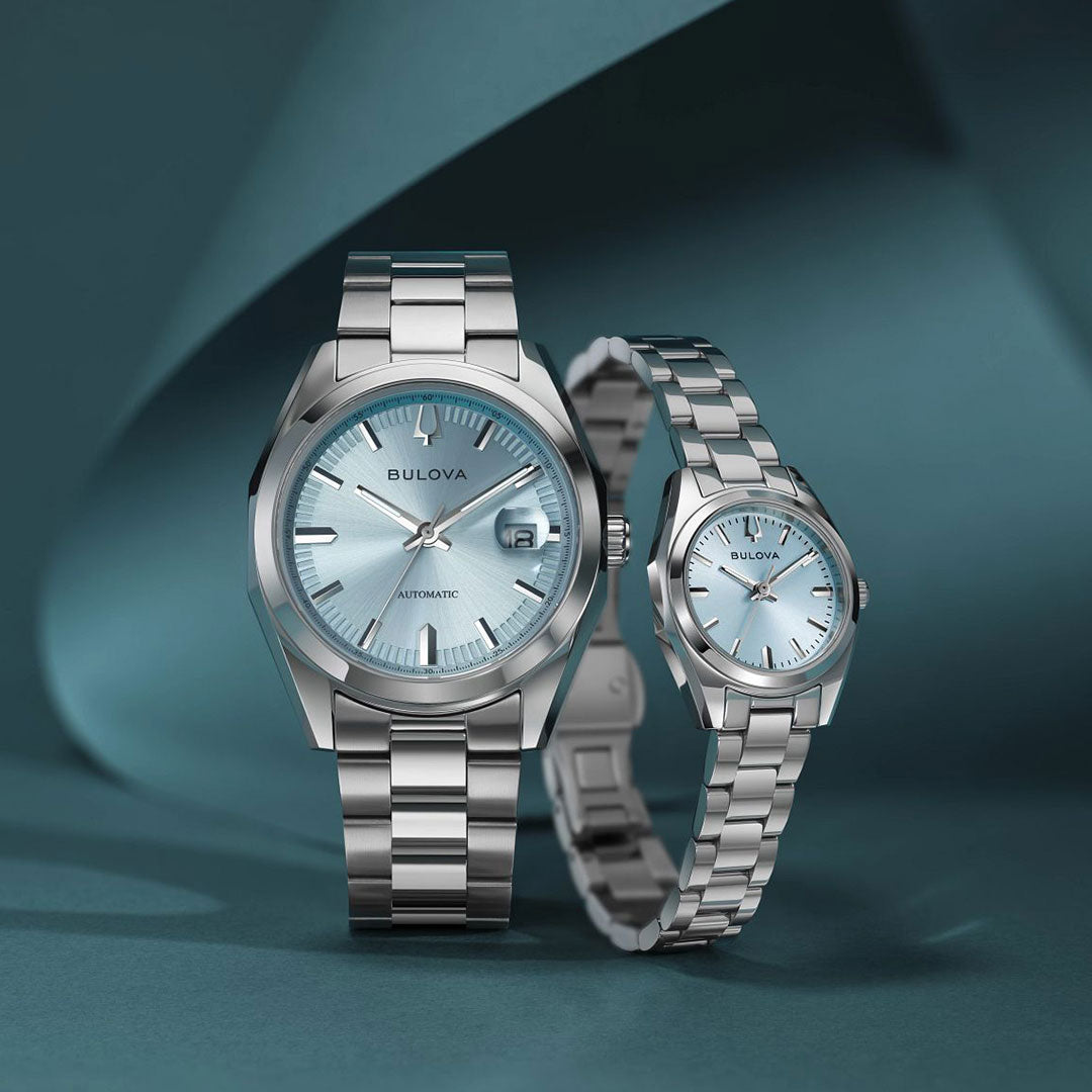 Two Bulova Surveyor Automatic 39mm watches with stainless steel bands and light blue dials are displayed on a wavy blue background. The men’s watch is on the left, the smaller version on the right—both feature elegant automatic movement.