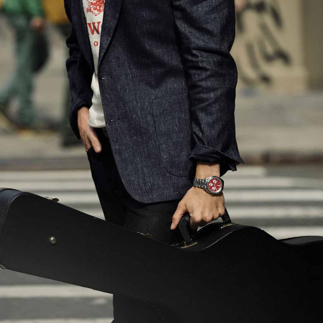 A person in a dark blazer stands on a city street, firmly gripping a guitar case. On their wrist, the distinctive red face of the Bulova Lunar Pilot Quartz 43.5mm Watch gleams, showcasing its precision quartz movement with timeless elegance.