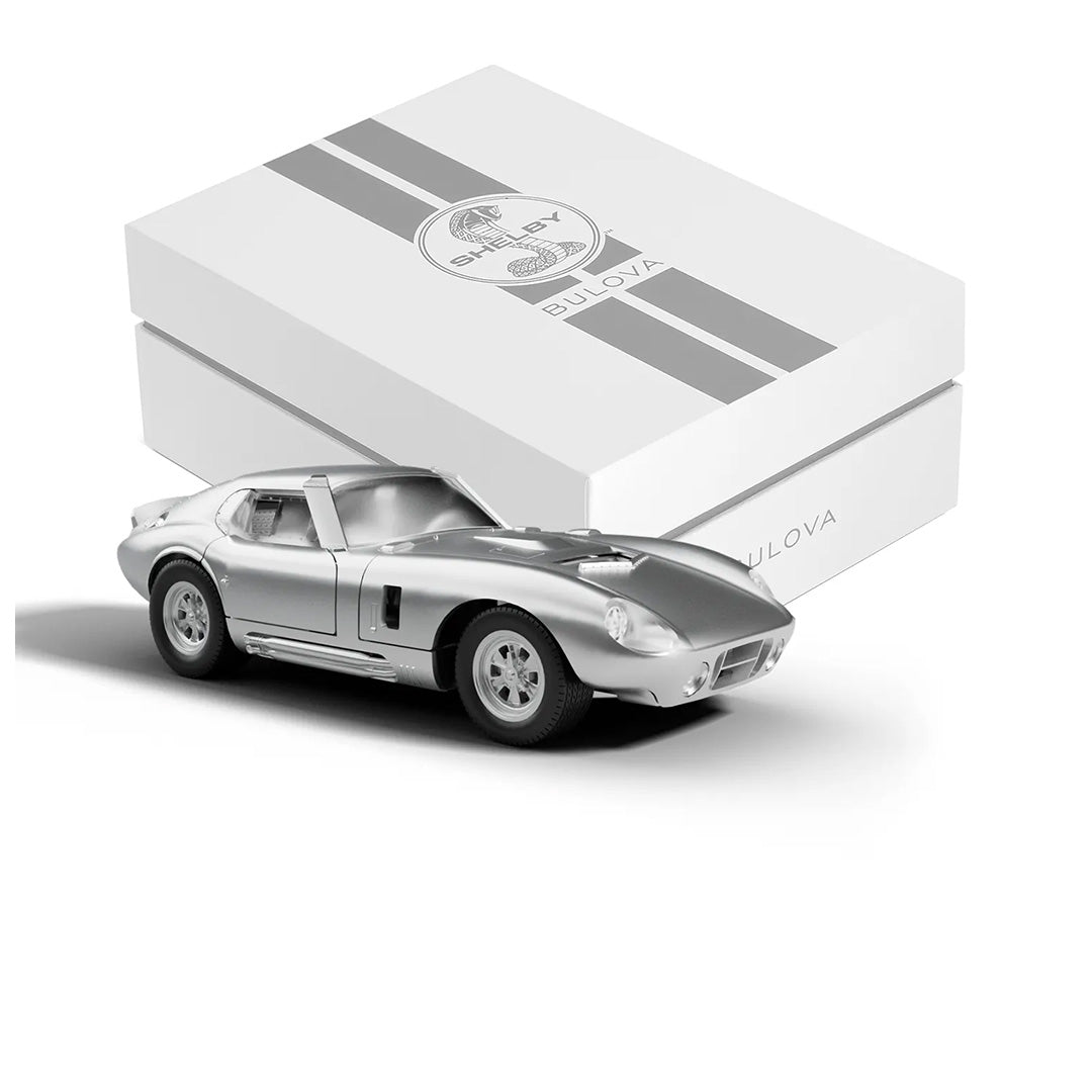 A silver model car is showcased in front of a white box with gray stripes featuring the Shelby and Bulova logos, perfectly embodying the essence of the Bulova Shelby Racer Chronograph Quartz 40.5mm Watch Limited Edition—an exceptional collectible for any automotive enthusiast and watch collector.