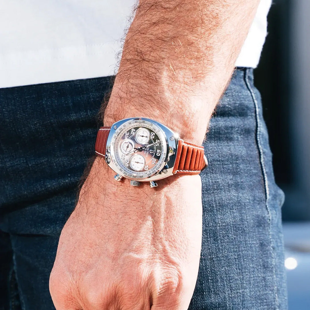 A close-up of a person’s wrist wearing the Bulova Shelby Racer Chronograph Quartz 40.5mm Watch Limited Edition, featuring a silver case and brown leather strap, paired with blue jeans and a white shirt.
