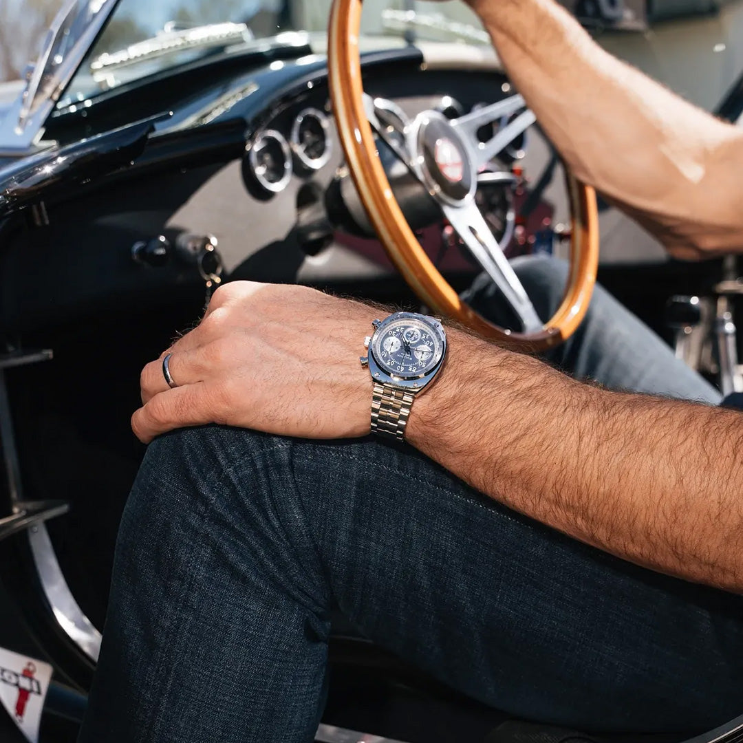 A person wearing a Bulova Shelby Racer Chronograph Quartz 40.5mm Watch Limited Edition and a wedding ring sits in a classic car, holding the wooden steering wheel. The focus is on their hand, the Bulova automotive enthusiast watch, and part of the car’s interior.