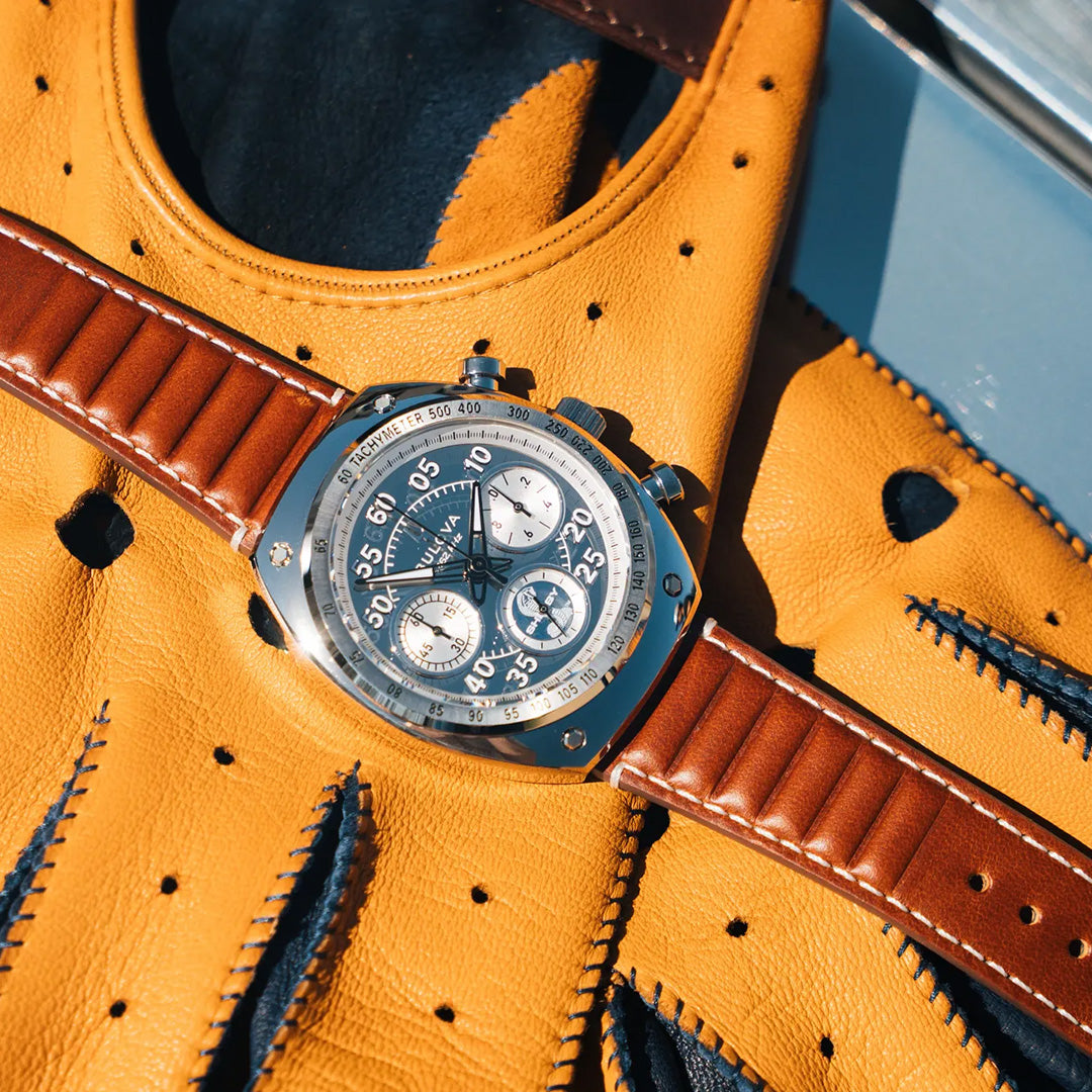 A Bulova Shelby Racer Chronograph Quartz 40.5mm Watch Limited Edition by Bulova, featuring a brown leather strap and silver chronograph design, rests on a yellow and black perforated driving glove in bright sunlight—making it the perfect timepiece for automotive enthusiasts.