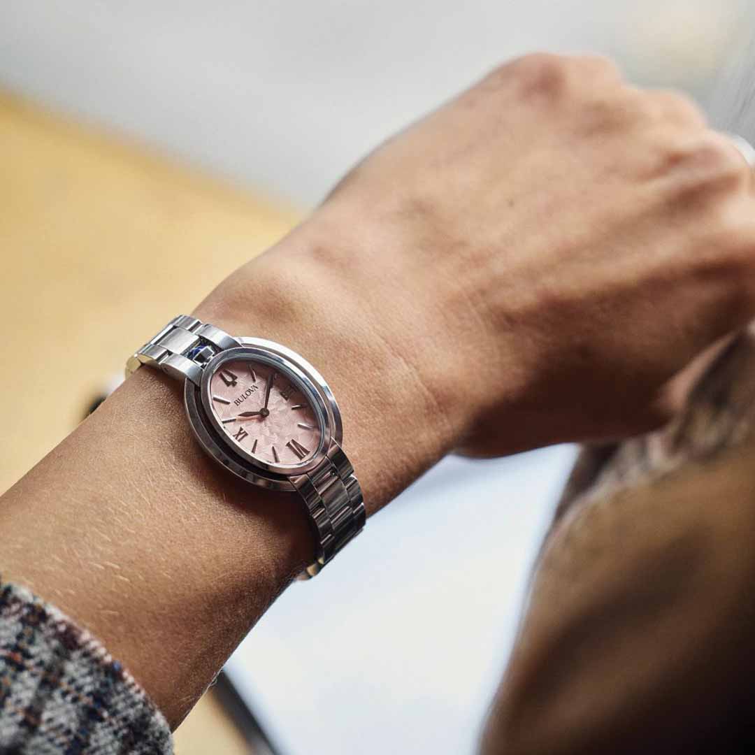 Close-up of a wrist adorned with a Bulova Rubaiyat Quartz 28.8mm Watch, showcasing a silver timepiece with a pink face. The softly blurred background highlights the watch, which rests gracefully on a stainless steel bracelet, displaying the time as 10:09.