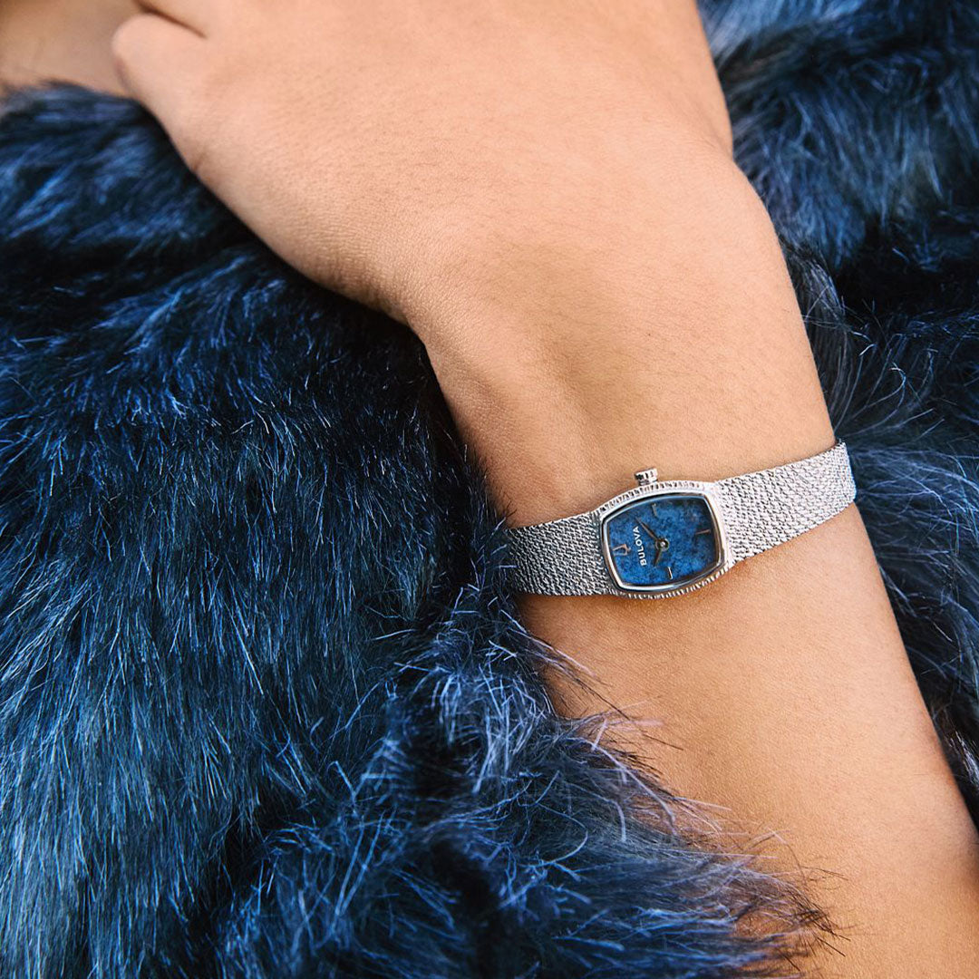A hand wearing the Bulova Goddess of Time Quartz 17mm watch rests on a dark blue faux fur garment.