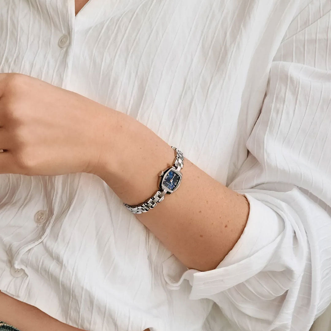 A person dressed in a white, textured shirt wears the Bulova Goddess of Time Jackie Quartz 18mm Watch on their left wrist. The watch, from Bulova's Goddess of Time collection, features a striking blue dial. Their arm is bent and partially raised.