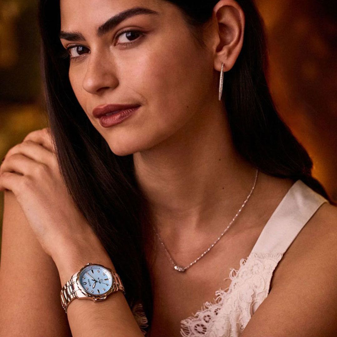 A woman with long dark hair wears a sleeveless white dress, hoop earrings, and the Bulova Marine Star Quartz 36mm Watch. Her delicate necklace complements this elegant timepiece gracing her wrist as she gazes softly at the camera against a warm, blurred background.