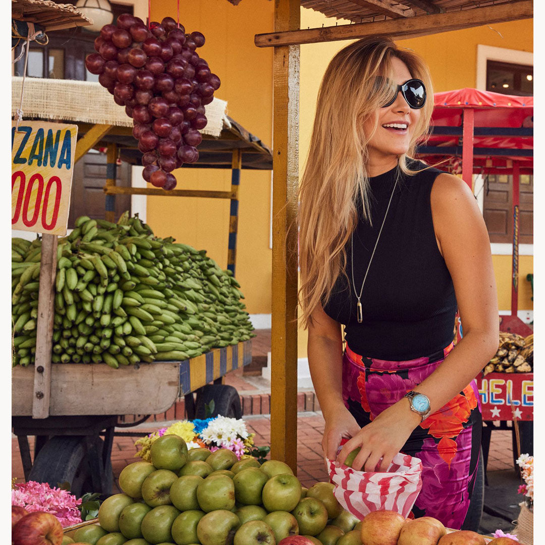 A woman with long blonde hair wearing sunglasses and a black sleeveless top is smiling at an outdoor market. Her wrist subtly glimmers with the Bulova Marine Star Quartz 36mm Watch, featuring diamond hour markers and a mother-of-pearl dial. She stands near stalls of bananas, grapes, and apples against colorful building backdrops.