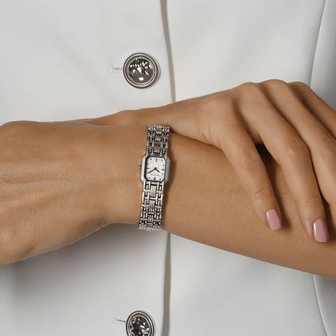 A person dressed in a white outfit with metallic buttons displays the Bulova Goddess of Time Sophia Quartz 16mm Watch on their left wrist. The rectangular face and woven band of this Bulova timepiece are elegantly complemented by their light pink nail polish.
