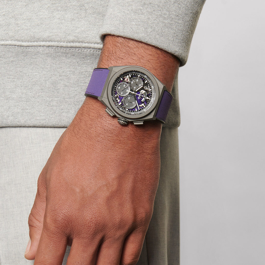 A person wearing a gray sweatshirt and light gray pants displays a ZENITH Defy El Primero 21 watch on their wrist. This intricate Zenith chronograph features a purple strap and a detailed, partially transparent face, revealing gears and small dials in matte titanium.