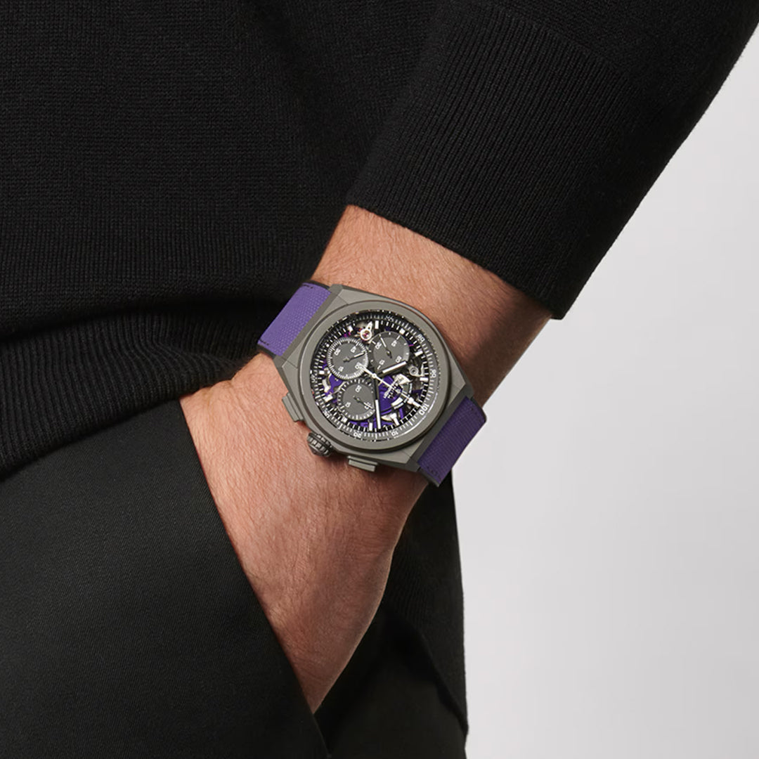 A close-up shows a wrist adorned with the ZENITH Defy El Primero 21 44mm Watch featuring a purple strap. The chronograph's intricate dial is set in matte titanium, revealing various mechanisms against a sleek, dark sleeve.