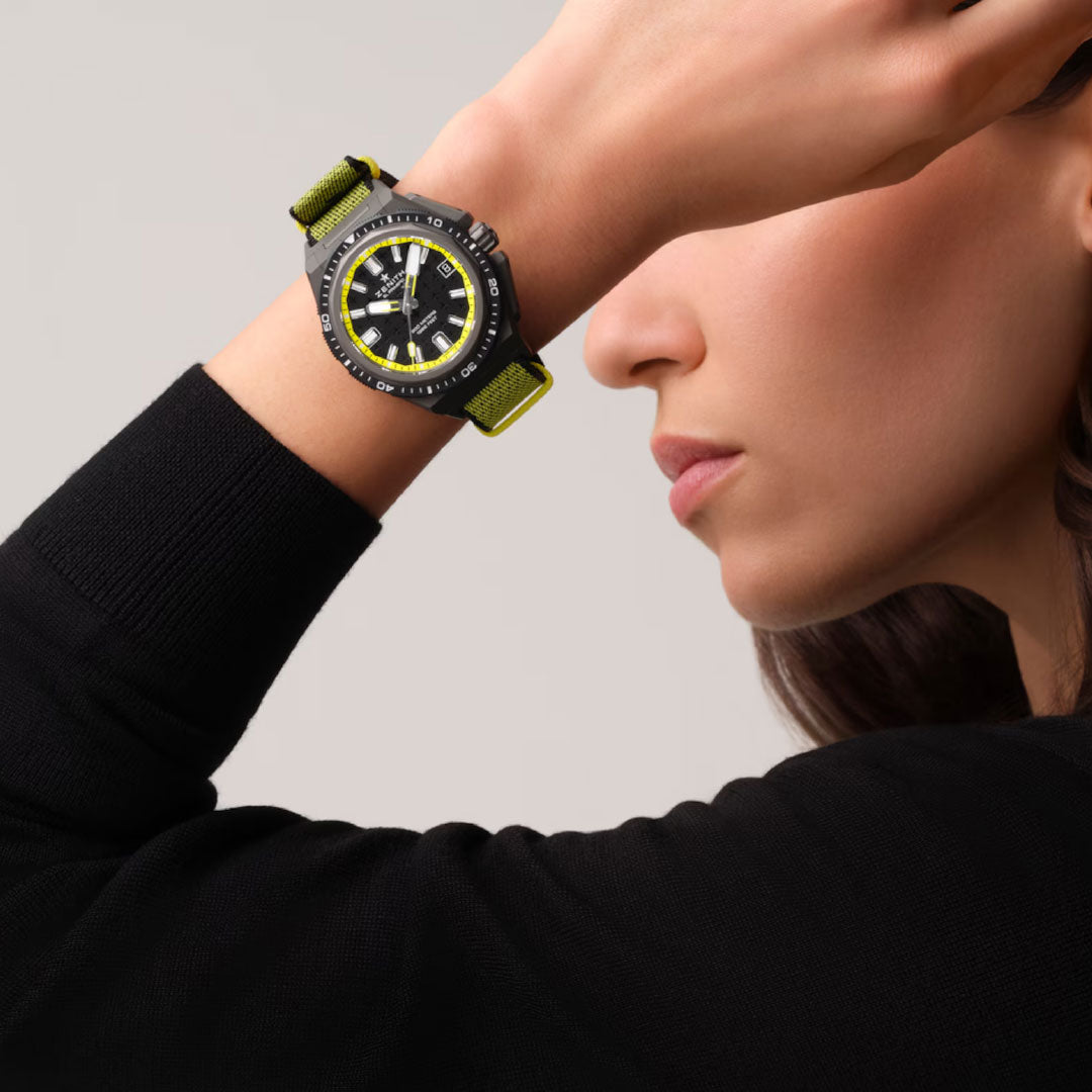 A woman in a black top poses with her arm raised, showcasing a Zenith Defy Extreme Diver El Primero 3620 43mm watch featuring a yellow and black strap and yellow dial accents, set against a neutral background.